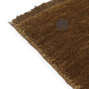 May include: Close-up of a brown rug with a textured surface and fringed edges. The rug is a solid color and appears to be made of a natural fiber. The corner of the rug is visible against a white background.