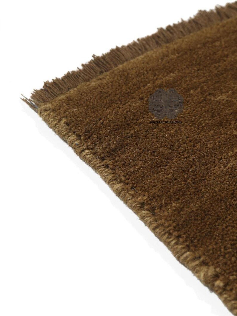 May include: Close-up of a brown rug with a textured surface and fringed edges. The rug is a solid color and appears to be made of a natural fiber. The corner of the rug is visible against a white background.