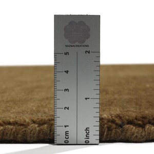 May include: Close-up of a brown rug with a ruler showing a thickness of approximately 1 inch. The ruler is marked with both inches and centimeters. The rug has a textured surface and a visible edge.