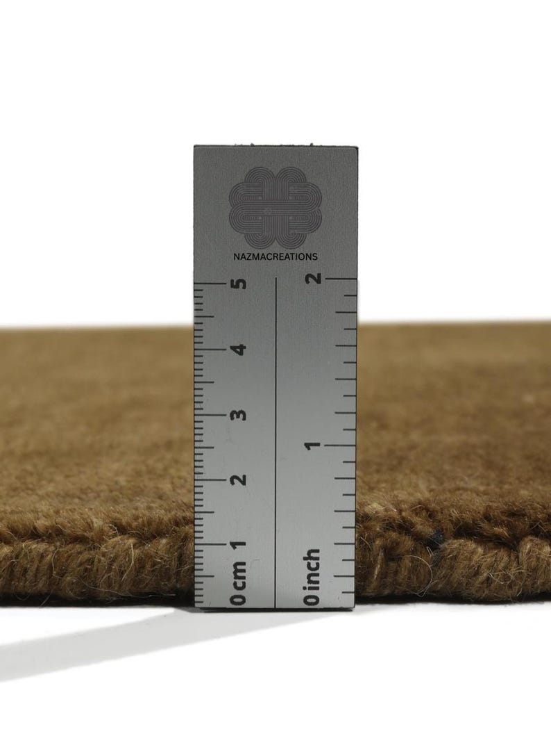 May include: Close-up of a brown rug with a ruler showing a thickness of approximately 1 inch. The ruler is marked with both inches and centimeters. The rug has a textured surface and a visible edge.
