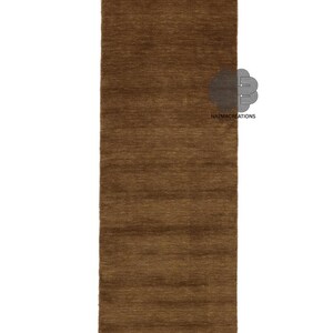 May include: A rectangular, brown rug with a textured surface. The rug is a solid color and appears to be made of a woven material. The rug is displayed on a white background. The logo "NAZMACREATIONS" is visible on the rug.