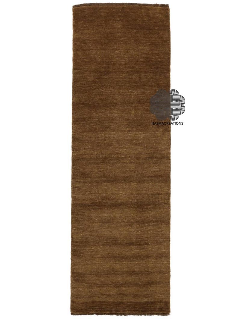 May include: A rectangular, brown rug with a textured surface. The rug is a solid color and appears to be made of a woven material. The rug is displayed on a white background. The logo "NAZMACREATIONS" is visible on the rug.