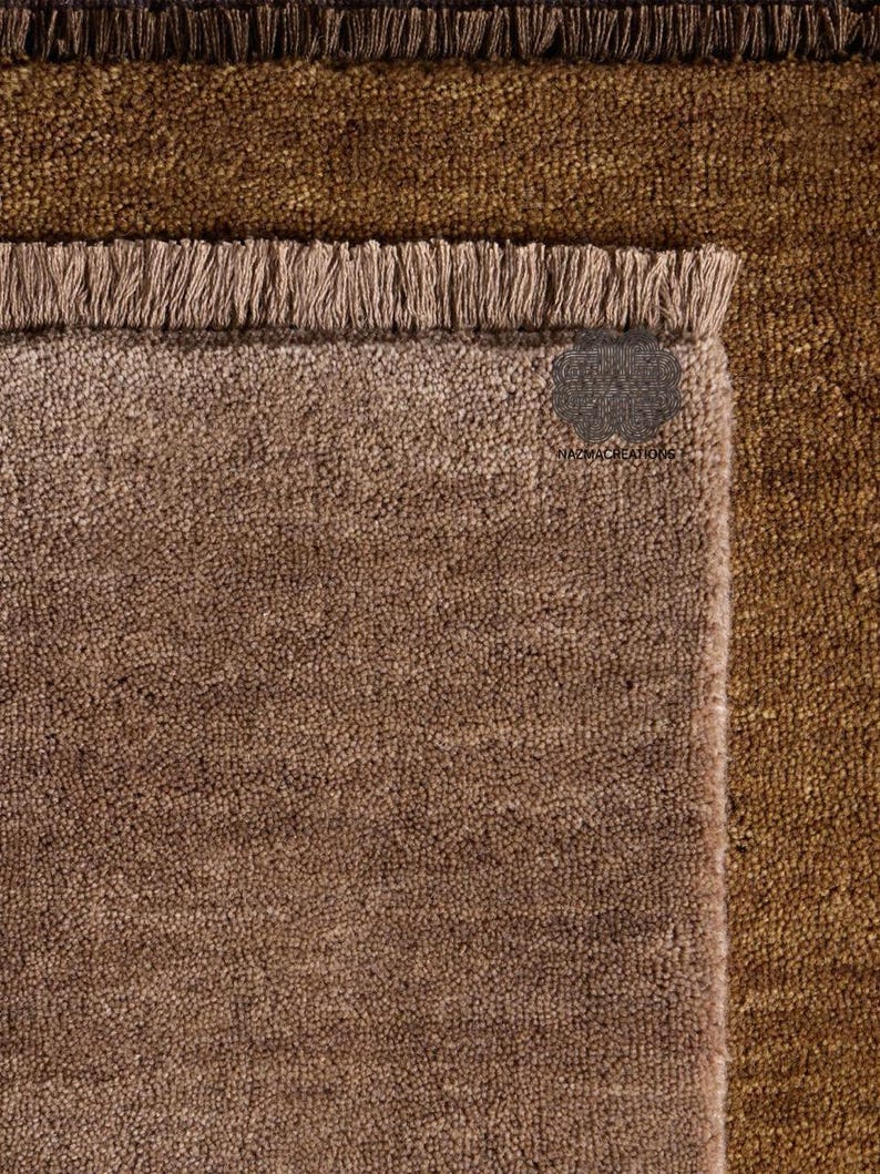 May include: Close-up of three textured rugs in shades of brown. The top rug is a dark brown, the middle rug is a lighter brown, and the bottom rug is a medium brown. Each rug has a fringed edge. The tag on the bottom rug reads "NAZMACREATIONS".