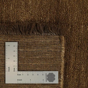 May include: Close-up of a brown woven rug with a textured surface and fringe detail. A ruler is placed on the rug, showing measurements in inches and centimeters. The rug has a natural, earthy appearance.
