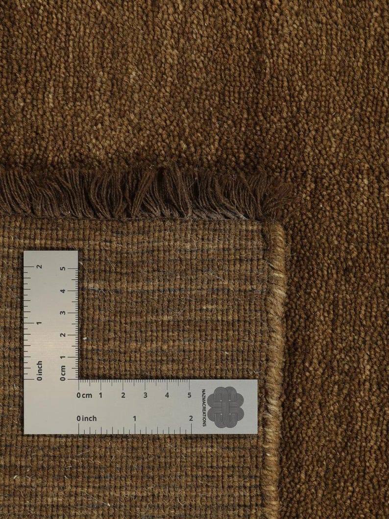 May include: Close-up of a brown woven rug with a textured surface and fringe detail. A ruler is placed on the rug, showing measurements in inches and centimeters. The rug has a natural, earthy appearance.