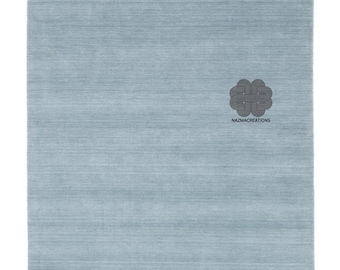 May include: A rectangular, light blue rug with a textured surface. The rug has a darker blue border and a logo in the bottom right corner that reads "NAZMACREATIONS". The rug is suitable for a living room or bedroom.