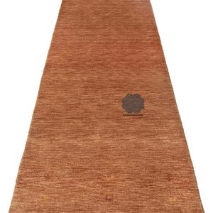 May include: A rectangular, rust-colored rug with a textured surface. The rug features a subtle pattern and a dark floral emblem near the center. The text "NAZMACREATIONS" is below the emblem. This rug is suitable for various interior settings.