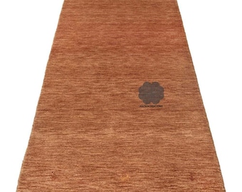 May include: A rectangular, rust-colored rug with a textured surface. The rug features a subtle pattern and a dark floral emblem near the center. The text "NAZMACREATIONS" is below the emblem. This rug is suitable for various interior settings.