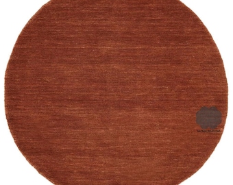 May include: A round, rust-colored rug with a textured surface. The rug is a solid color and appears to be made of woven material. The rug has a logo in the bottom right corner.
