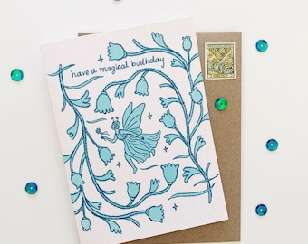 May include: A birthday card with a teal illustration of a fairy and floral elements. The card reads "have a magical birthday". The card is on a brown envelope with a decorative stamp and surrounded by teal sequins.