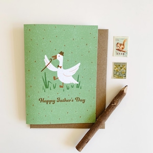 May include: A Father's Day greeting card with a green background and a cartoon goose wearing a hat and bow tie, holding a cane. The card reads "Happy Father's Day." Includes a brown envelope, stamps, and a wooden pencil.