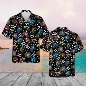 Op de afbeelding: Zwart overhemd met een patroon van kleurrijke monstertrucks, sterren en de tekst "CUSTOM NAME". Het shirt met korte mouwen heeft een witte kraag en wordt getoond op een houten oppervlak met een strandachtergrond.