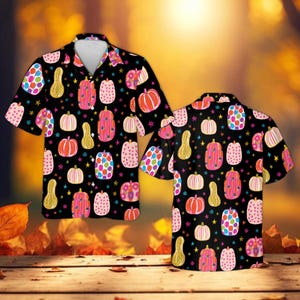 Può includere: Una camicia a maniche corte con bottoni su sfondo nero caratterizzata da un motivo ripetitivo di zucche e zucche ornamentali colorate nei toni del rosa, arancione e blu, intervallate da piccole stelle gialle su tutto il tessuto.