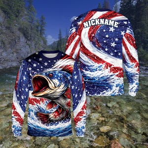 May include: Long-sleeved shirt with a patriotic design, featuring a large fish illustration. The shirt's design incorporates the American flag's stars and stripes, with the word "NICKNAME" on the back. The fish is depicted with an open mouth, set against a wave background.