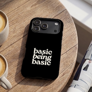 May include: A black smartphone case featuring the white text "basic being basic" in a bold, serif font. The case has a rectangular shape with a cutout for a triple-lens camera system on the back, showcasing a clean and simple design aesthetic.