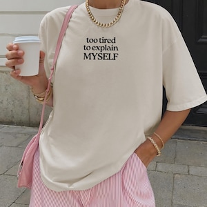 May include: An off-white short-sleeve t-shirt featuring the black text "too tired to explain MYSELF" printed on the chest. The shirt is paired with pink and white pinstriped shorts and a pink shoulder bag, accessorized with a gold chain necklace.