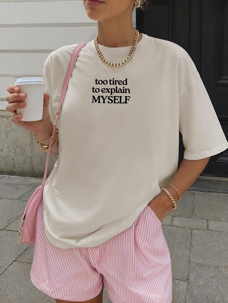 May include: An off-white short-sleeve t-shirt featuring the black text "too tired to explain MYSELF" printed on the chest. The shirt is paired with pink and white pinstriped shorts and a pink shoulder bag, accessorized with a gold chain necklace.
