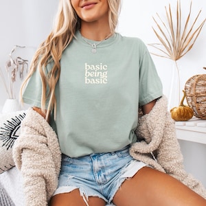 Puede incluir: Camiseta verde salvia con el texto blanco "basic being basic". La camiseta se combina con pantalones cortos de mezclilla y una rebeca color crema. En el fondo hay calabazas decorativas y una mesa blanca.