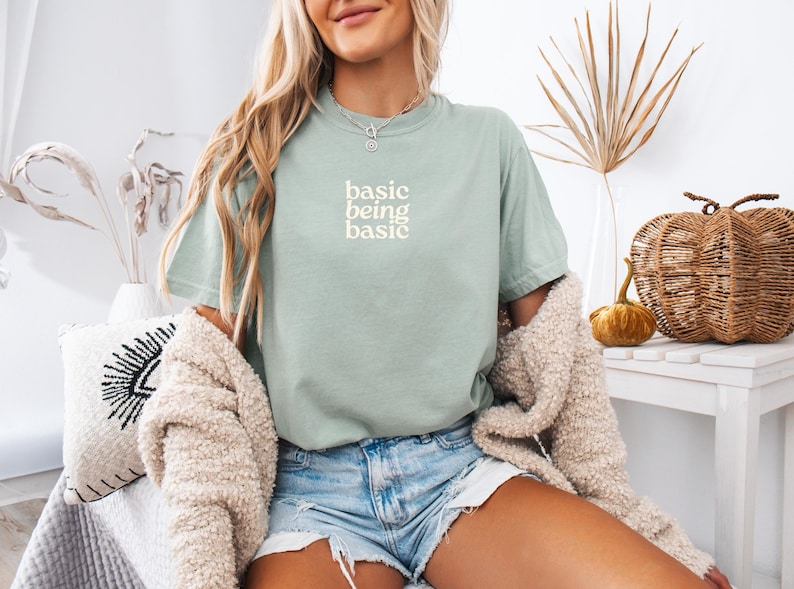 Puede incluir: Camiseta verde salvia con el texto blanco "basic being basic". La camiseta se combina con pantalones cortos de mezclilla y una rebeca color crema. En el fondo hay calabazas decorativas y una mesa blanca.