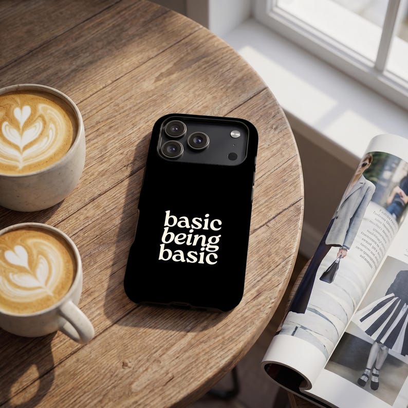 Black iPhone case with white “basic being basic” text, shown on a wooden coffee table with latte cups and magazine, DJO Basic Being Basic phone case mockup.