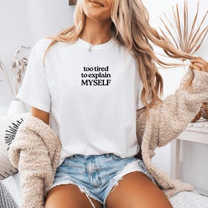 May include: A white short-sleeved t-shirt featuring the black text "too tired to explain MYSELF" printed on the chest. The shirt is paired with light blue denim shorts and a beige textured cardigan, creating a casual layered look.