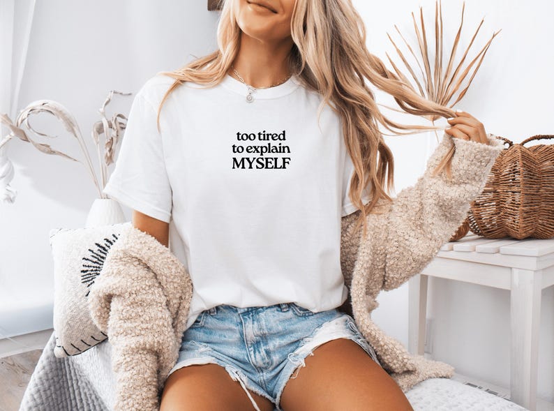 May include: A white short-sleeved t-shirt featuring the black text "too tired to explain MYSELF" printed on the chest. The shirt is paired with light blue denim shorts and a beige textured cardigan, creating a casual layered look.