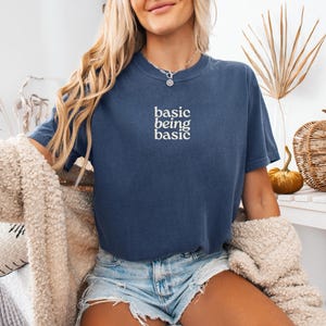 Puede incluir: Una camiseta azul marino con el texto blanco "basic being basic". La camiseta se usa con pantalones cortos de mezclilla y un c&aacute;rdigan color crema. Una calabaza decorativa y plantas secas est&aacute;n en el fondo.