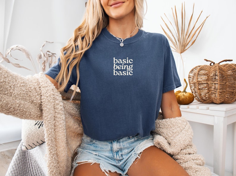 Puede incluir: Una camiseta azul marino con el texto blanco "basic being basic". La camiseta se usa con pantalones cortos de mezclilla y un c&aacute;rdigan color crema. Una calabaza decorativa y plantas secas est&aacute;n en el fondo.