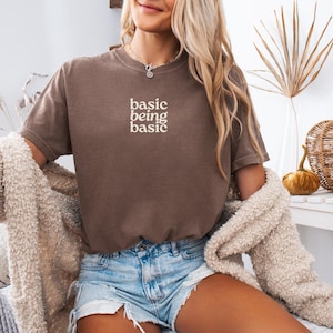 Puede incluir: Una camiseta marr&oacute;n con el texto "basic being basic" en blanco. La modelo lleva pantalones cortos vaqueros y una rebeca color crema. En el fondo hay calabazas decorativas y una calabaza tejida.