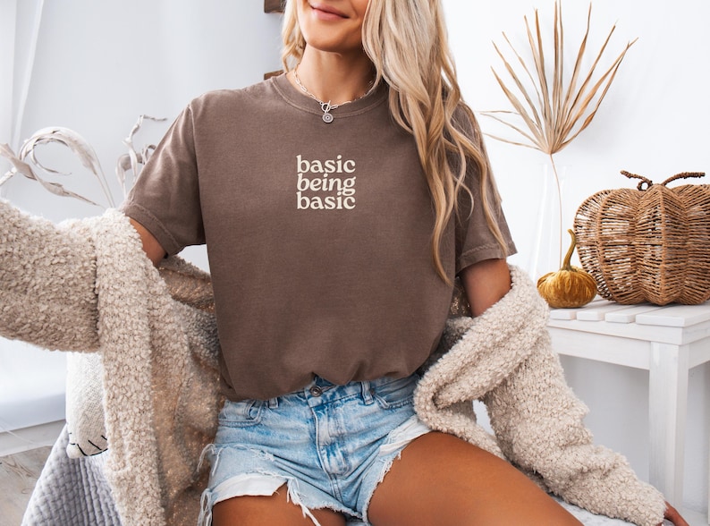 Puede incluir: Una camiseta marr&oacute;n con el texto "basic being basic" en blanco. La modelo lleva pantalones cortos vaqueros y una rebeca color crema. En el fondo hay calabazas decorativas y una calabaza tejida.