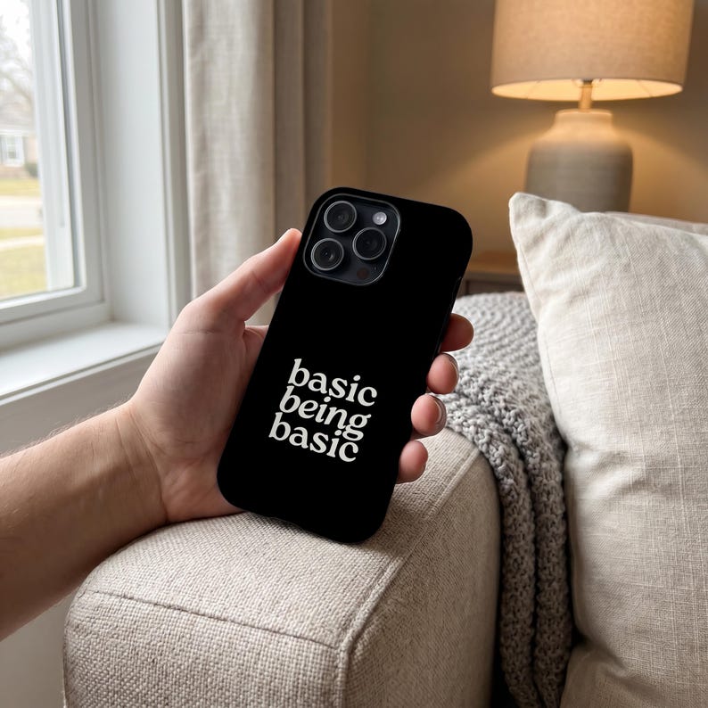 May include: A black smartphone case featuring the white text "basic being basic" printed in a stylized serif font. The case has a cutout for a triple-lens camera system on the back and covers the rear and sides of the mobile device.