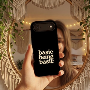 May include: A black smartphone case featuring the text "basic being basic" printed in a cream-colored, sans-serif font. The case has a cutout for the camera lens and flash at the top, with a smooth, matte finish covering the back of the device.