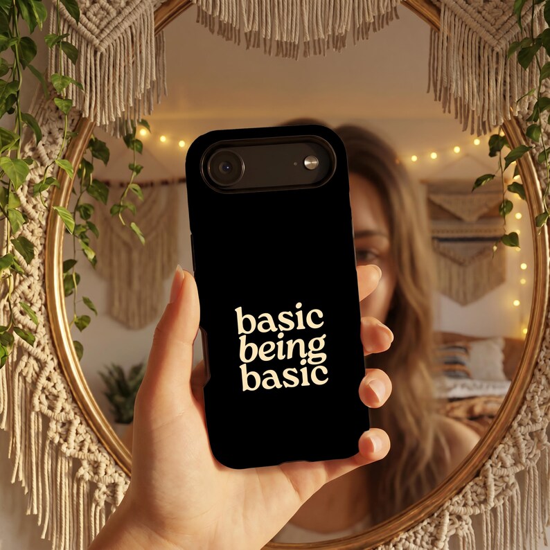 May include: A black smartphone case featuring the text "basic being basic" printed in a cream-colored, sans-serif font. The case has a cutout for the camera lens and flash at the top, with a smooth, matte finish covering the back of the device.