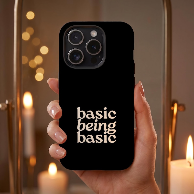 Black Basic Being Basic iPhone case with white text, held in hand against warm candlelight for a cozy evening lifestyle mockup.