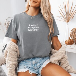 May include: A slate gray short-sleeve t-shirt featuring the white printed text "too tired to explain MYSELF" centered on the chest. The garment has a crew neckline and a standard fit, paired with light blue denim shorts and a textured beige cardigan.