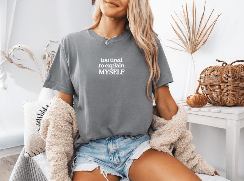 May include: A slate gray short-sleeve t-shirt featuring the white printed text "too tired to explain MYSELF" centered on the chest. The garment has a crew neckline and a standard fit, paired with light blue denim shorts and a textured beige cardigan.