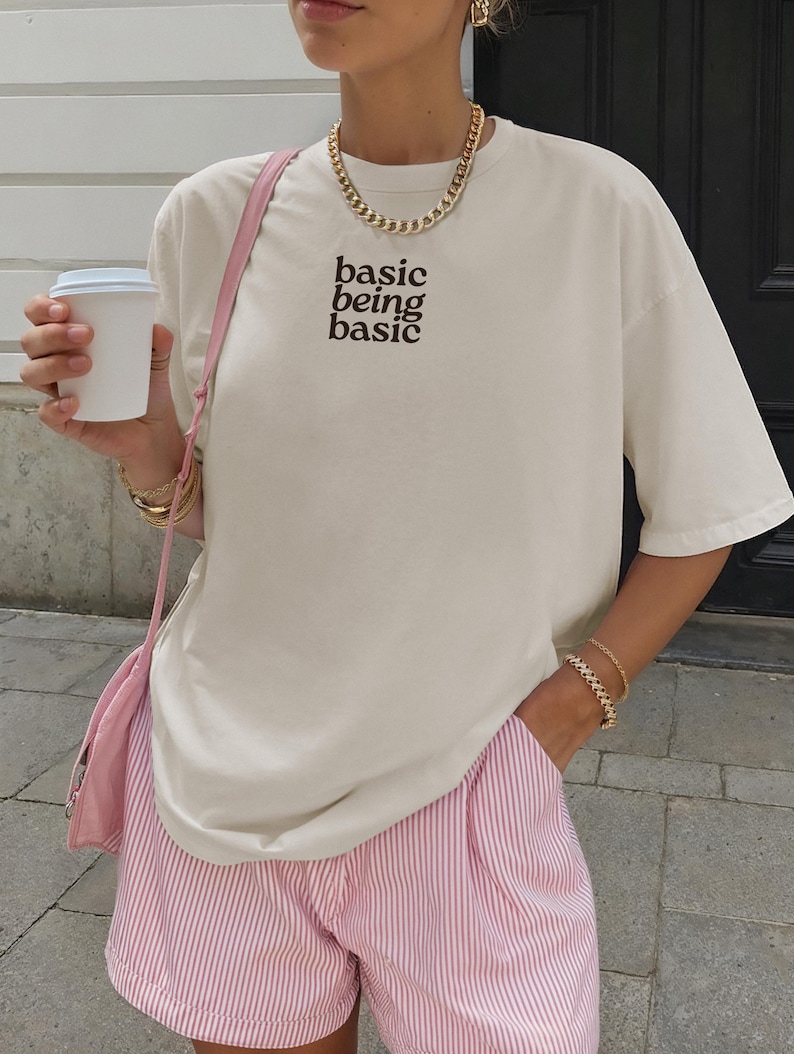Puede incluir: Camiseta color crema con el texto "basic being basic" en marr&oacute;n. La camiseta se combina con shorts a rayas rosas y blancas. Se lleva un collar y pulseras de cadena dorada. Tambi&eacute;n se ven un bolso de hombro rosa y una taza de caf&eacute; blanca.