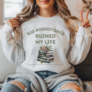 Może przedstawiać: Biała bluza z napisem "BIG BOOKSTORES RUINED MY LIFE" w kolorze zielonym. Poniżej tekstu znajduje się stos książek z kwiatowymi akcentami. Bluza z długim rękawem jest noszona przez osobę.
