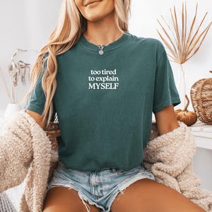 May include: A dark teal short-sleeve t-shirt featuring the white text "too tired to explain MYSELF" printed on the chest. The shirt has a crew neckline and a relaxed fit, paired with light blue denim shorts and a textured beige cardigan.