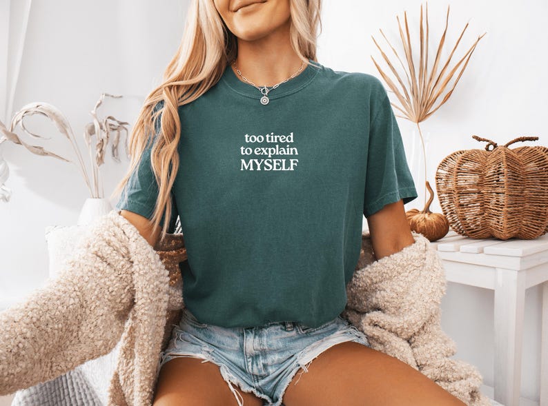 May include: A dark teal short-sleeve t-shirt featuring the white text "too tired to explain MYSELF" printed on the chest. The shirt has a crew neckline and a relaxed fit, paired with light blue denim shorts and a textured beige cardigan.