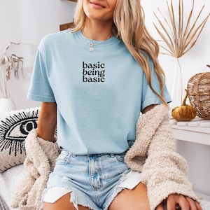 Puede incluir: Camiseta azul claro con el texto negro "basic being basic". La camiseta se usa con pantalones cortos de mezclilla y una rebeca color crema. La imagen est&aacute; decorada con calabazas decorativas y un coj&iacute;n con un dise&ntilde;o de ojo.