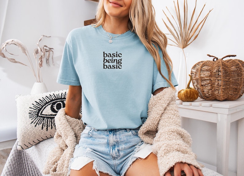 Puede incluir: Camiseta azul claro con el texto negro "basic being basic". La camiseta se usa con pantalones cortos de mezclilla y una rebeca color crema. La imagen est&aacute; decorada con calabazas decorativas y un coj&iacute;n con un dise&ntilde;o de ojo.