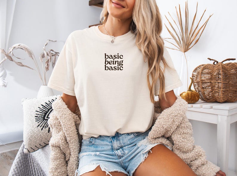 May include: Cream-colored t-shirt with the text "basic being basic" in a black, minimalist font. The shirt is paired with denim shorts. The overall aesthetic is casual and relaxed, suitable for everyday wear.