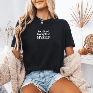 May include: A black short-sleeved t-shirt featuring the white text "too tired to explain MYSELF" printed on the center of the chest. The shirt has a crew neckline and is paired with light blue denim shorts and a beige textured cardigan.