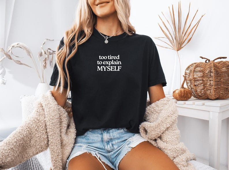 May include: A black short-sleeved t-shirt featuring the white text "too tired to explain MYSELF" printed on the center of the chest. The shirt has a crew neckline and is paired with light blue denim shorts and a beige textured cardigan.