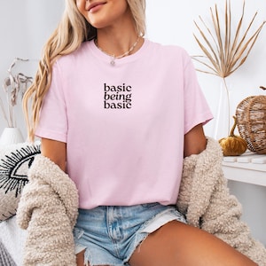 Puede incluir: Camiseta rosa claro con la frase "basic being basic" en marr&oacute;n oscuro. La camiseta de manga corta tiene cuello redondo. La persona lleva pantalones cortos vaqueros y una rebeca beige. Tema: ropa informal.