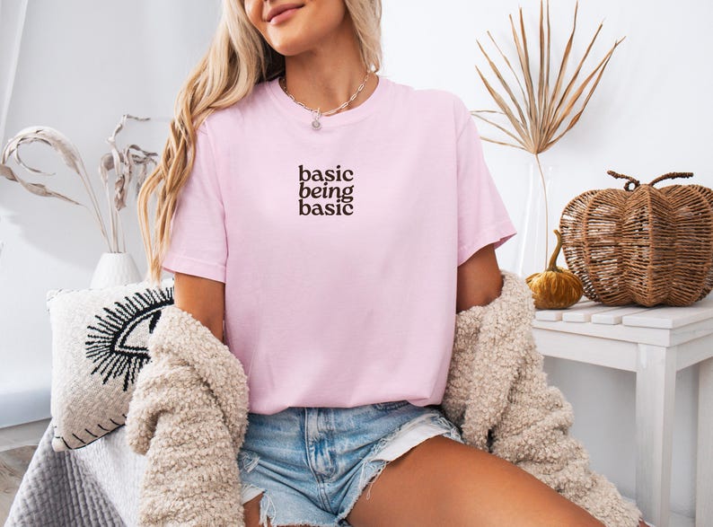 Puede incluir: Camiseta rosa claro con la frase "basic being basic" en marr&oacute;n oscuro. La camiseta de manga corta tiene cuello redondo. La persona lleva pantalones cortos vaqueros y una rebeca beige. Tema: ropa informal.
