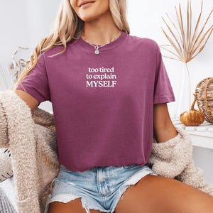 May include: A plum-colored short-sleeve t-shirt featuring the white text "too tired to explain MYSELF" printed on the chest. The shirt has a crew neckline and a relaxed fit, paired with light blue denim shorts and a textured beige cardigan.