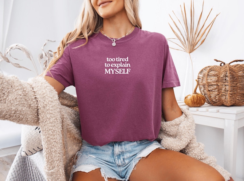 May include: A plum-colored short-sleeve t-shirt featuring the white text "too tired to explain MYSELF" printed on the chest. The shirt has a crew neckline and a relaxed fit, paired with light blue denim shorts and a textured beige cardigan.