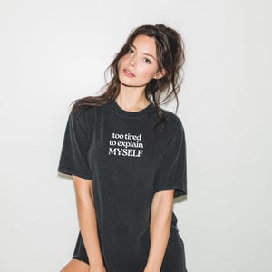 May include: A black short-sleeved t-shirt featuring the white printed text "too tired to explain MYSELF" centered on the chest. The garment has a crew neckline and a relaxed fit, constructed from a dark, slightly faded black cotton fabric.